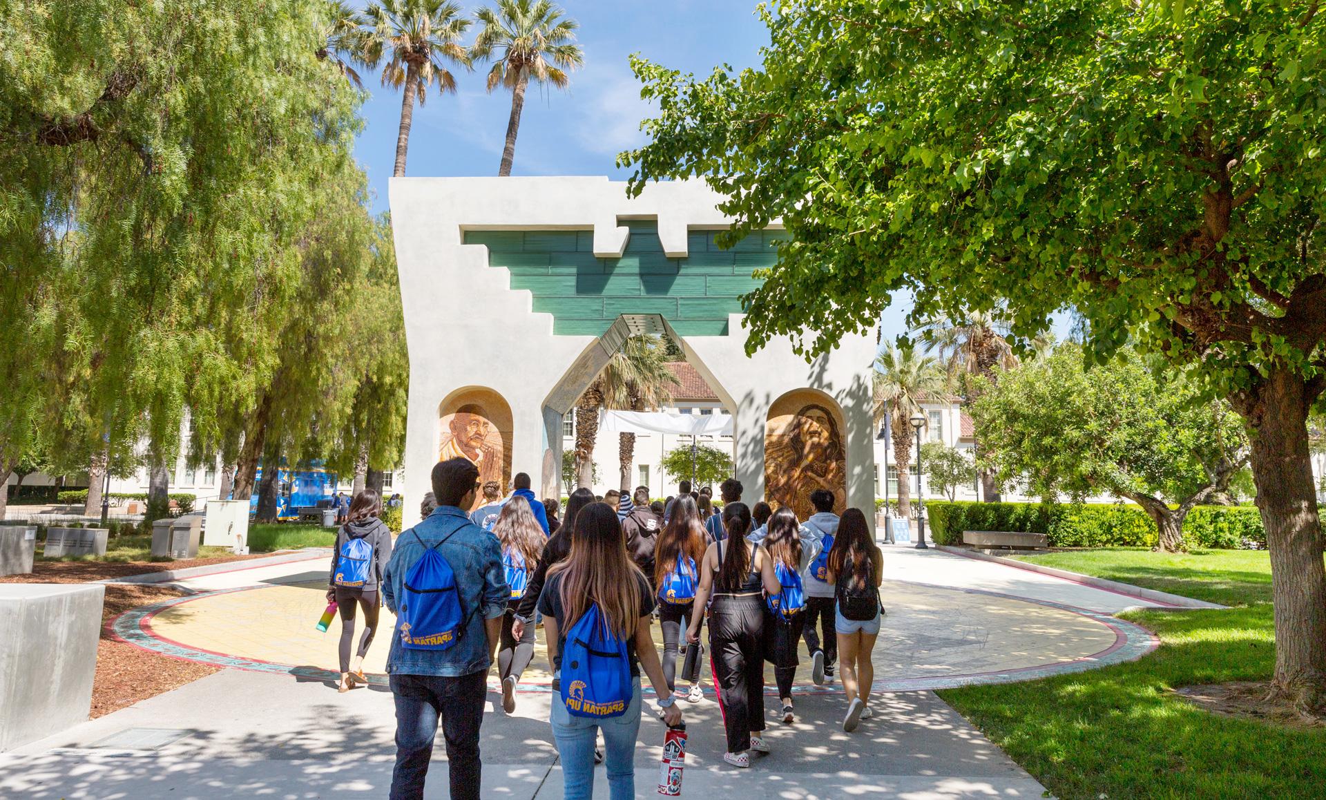 Students wearing 十大菠菜软件 gear are walking through the Cesar Chavez arch that is surrounded by bright green trees.